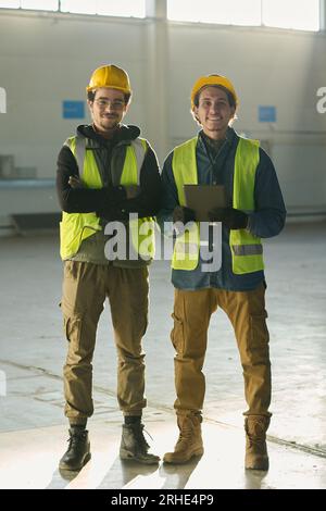 Happy young male colleagues in safety helmets and reflective jackets standing in front of camera in workshop while one of them using tablet Stock Photo