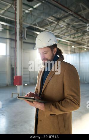 Young serious contractor looking through online information on tablet screen while standing in front of camera at construction site Stock Photo