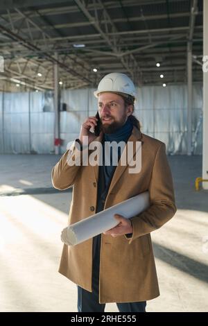Young bearded engineer with smartphone by ear and rolled blueprint in hand standing in front of camera in warehouse and calling someone Stock Photo