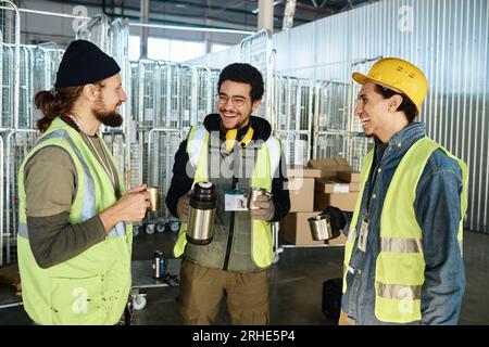 Three young cheerful engineers in workwear looking at each other while discussing something funny and having hot tea at break in warehouse Stock Photo