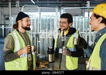 Three happy young workers with metallic mugs of hot tea having chat while standing in front of camera against large carts in warehouse Stock Photo