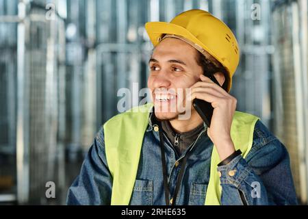 Young smiling multiethnic male engineer in safety helmet talking on the phone to colleague or his family while sitting in front of camera at break Stock Photo