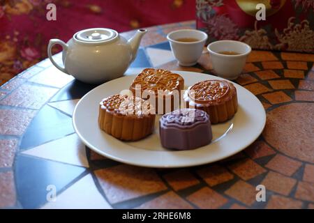Four different Chinese Mooncakes on a white plate, round, square biscuit-coloured pastry and a purple hexagonal cake,  two teacups and a teapot. Stock Photo