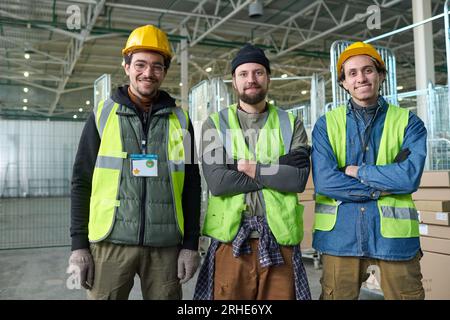 Group of happy young intercultural workers of warehouse or factory in safety helmets and fluorescent jackets standing in front of camera Stock Photo