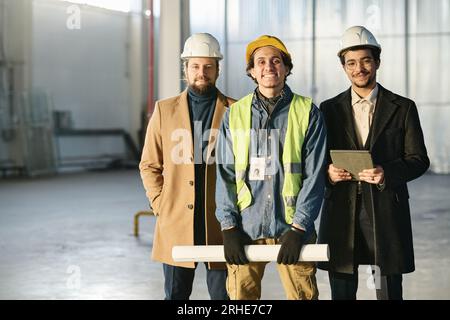 Group of happy young intercultural men in hardhats looking at camera while standing against wall and corner of spacious warehouse Stock Photo