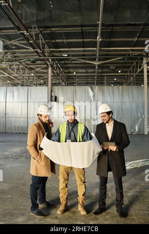 Front view of team of young architects or engineers discussing building plan or sketch on unrolled blueprint held by worker in safety vest Stock Photo