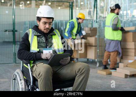 Young serious worker of warehouse sitting in wheelchair in front of camera against two colleagues loading boxes on carts and using tablet Stock Photo
