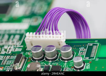 Electrolytic capacitors and white plastic connector with purple cables, crystal or diode on green PCB. Printed circuit board and electronic components. Stock Photo