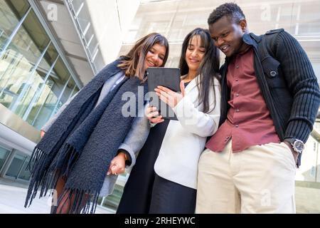 Smiling coworkers using a tablet at office outdoors. Group of young friends meeting in the city. Ethnic diversity. Stock Photo
