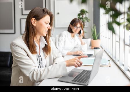 Female company employees working in office lady pointing at laptop screen while colleague talking on phone on background Stock Photo