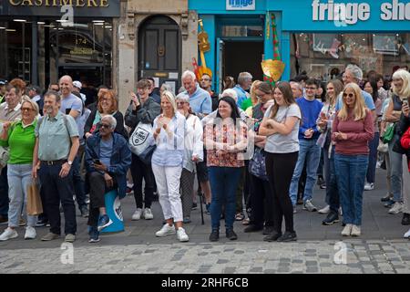 City centre, Edinburgh, Scotland, UK. 16 August 2023.  Cloudy conditions in the city centre for those visiting the Edinburgh Festival Fringe, small audiences for some street performers on the High Street. Pictured: A small audiience is entertained outside the Fring shop and office on the High Street.. Credit: Archwhite/alamy live news Stock Photo