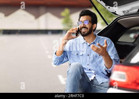 Excited guy sitting in open car trunk, have phone conversation Stock Photo