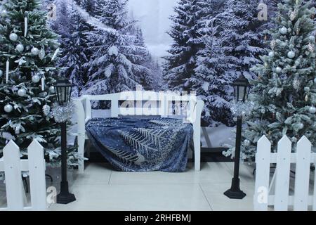 Photo zone: New Year's Christmas white bench on the background of blue, green fir trees, Christmas trees in the snow and a white fence. Festive mood. Stock Photo