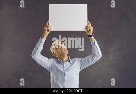 Caucasian senior man looks at white blank sheet of paper he holds over his head. Stock Photo
