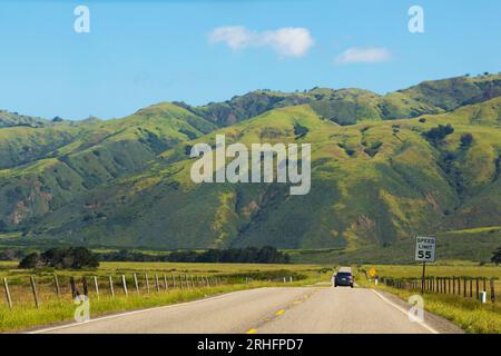 Road stretches into the distance against the backdrop of green meadows and mountains. Asphalt road with a dividing strip goes into the distance. Car drive far along the highway, rear view Stock Photo