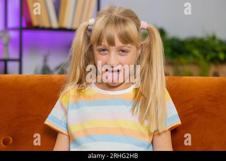 Caucasian joyful preteen school girl making playful silly facial expressions and grimacing, fooling around, showing tongue. Young excited blonde child kid at home room apartment sitting on couch Stock Photo