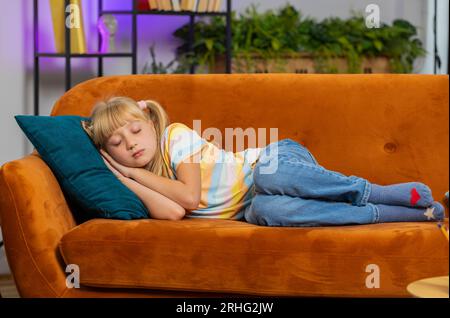 Tired preteen school girl lying down in bed taking a rest at home on couch. Carefree blonde child kid napping, falling asleep on comfortable sofa with pillows. Closed her eyes enjoy daytime nap alone Stock Photo