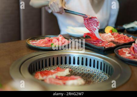 woman's hand employs tongs to place wagyu beef on a plate, ready to grill over charcoal, enhancing the dining experience at a Japanese restaurant Stock Photo