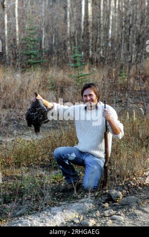 A vintage photograph from the year 1980 depicts a young male bird hunter proudly posing with a Hungarian partridge and a firearm in a forest. Stock Photo