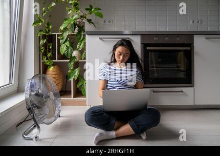 Handsome asian girl student with laptop is languishing heat sitting on cool floor next fan. Stock Photo