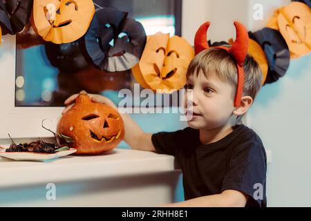 little caucasian 5 year old boy wearing devil horns sitting by Jack lantern made of orange pumpkin. Halloween concept. Image with selective focus. Stock Photo