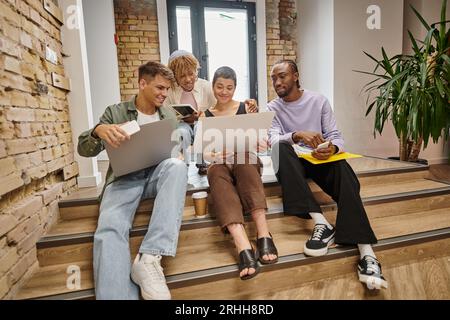 happy interracial team looking at laptop, holding gadgets, sitting on stairs in coworking, startup Stock Photo