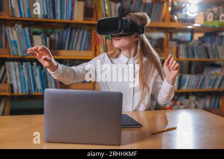 Girl student using modern tech devices in the library, a laptop and VR headset. Concepts of virtual reality experience in education. Stock Photo