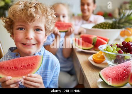 Smiling boy holding watermelon slice with fresh fruits on table in kitchen at home Stock Photo