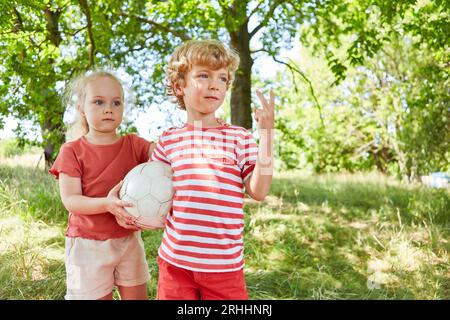 Brother showing peace sign while standing with sister holding soccer ball in garden Stock Photo