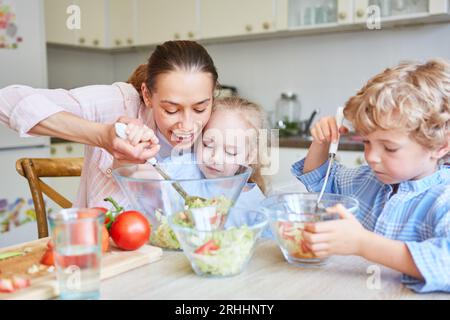 Mother helping daughter mixing salad with brother sitting in kitchen at home Stock Photo