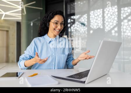 Young beautiful woman with headset phone talking to colleagues remotely using laptop for video call, latin american smiling joyfully looking at laptop web camera. Stock Photo