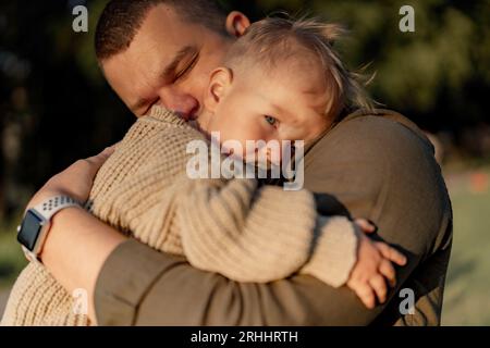 young dad with eyes closed hugging baby son putting head to his shoulder and sucking a pacifier. they are standing in park. High quality photo Stock Photo