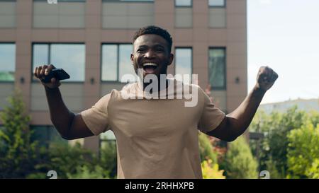 African American happy success winner man ethnic guy in city male outdoors win victory achieve with mobile phone smartphone achievement jump scream Stock Photo