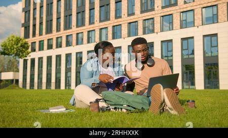 Two friends African American ethnic students sitting on green grass meadow lawn in park outdoors woman man girl guy doing homework university prepare Stock Photo