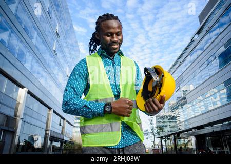 young civil engineer man of african ethnicity with beard and dreadlocks, holding his helmet with hand is standing outdoors smiling happy at end of wor Stock Photo