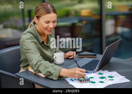 Smiling businesswoman working on laptop and making notes in cafe terrace while drinking coffee Stock Photo