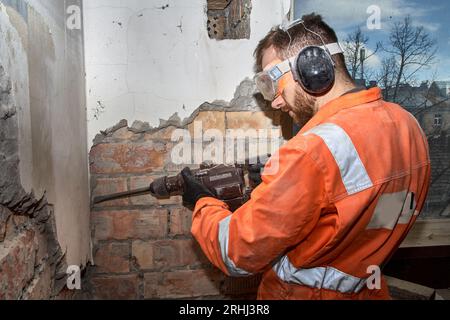 Construction worker using hammer drill with chisel for demolition jobs indoor, wearing orange coveralls, and PPE. Stock Photo