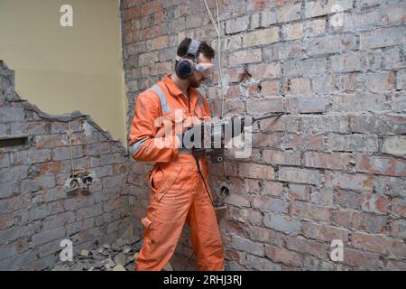 Construction worker using hammer drill with chisel to remove old cement from brick wall indoors, wearing gloves, mask, ear defenders and orange covera Stock Photo