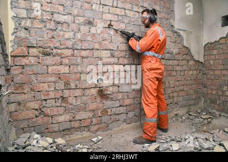 Worker standing removing old plaster from red brick wall by hammer drill, wearing gloves, boots, mask, ear defenders and orange coveralls. Stock Photo
