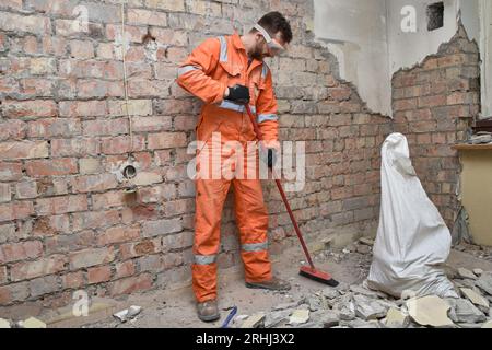 Builder cleaning room from old concrete and bricks after demolition jobs, wearing gloves, mask, boots and orange coveralls. Stock Photo