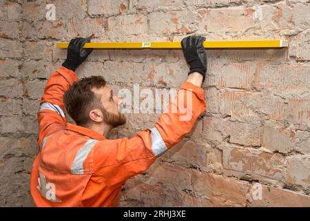 Young construction worker checking brick wall for level, wearing gloves and orange coveralls. Stock Photo