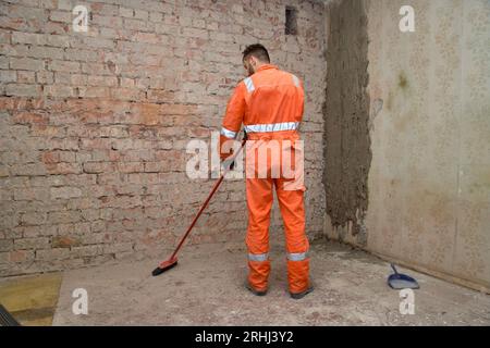 Builder cleaning room from dust after demolition jobs, wearing gloves, boots and orange coveralls. Stock Photo