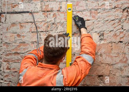 Construction worker drawing a vertical line on a brick wall indoor using a level, wearing gloves and orange coveralls. Stock Photo