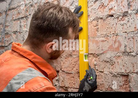 Builder drawing a vertical line on a brick wall using spirit level, wearing gloves and orange coveralls. Stock Photo