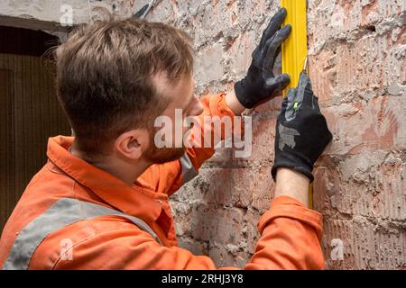 Young construction worker drawing a vertical line on a brick wall using spirit level, wearing gloves and orange coveralls. Stock Photo