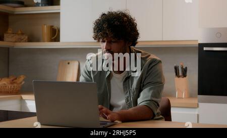 Concentrated Indian businessman typing computer working shopping online distant from home with laptop at kitchen serious focused freelancer Arabian Stock Photo