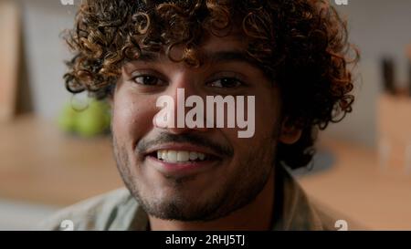 Close up smiling Arabian man homeowner with curly hair vlogger look at camera at kitchen. Portrait Indian male face with smile indoors. Headshot Stock Photo