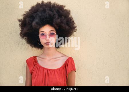 Portrait of serious young Moroccan female in sunglasses with Afro hair looking at camera while standing on street against beige wall in daylight Stock Photo