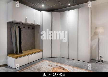Creative interior of minimal wardrobe with hanging clothes designed in angle among wooden cupboards with closed doors near carpet and floor lamp under Stock Photo