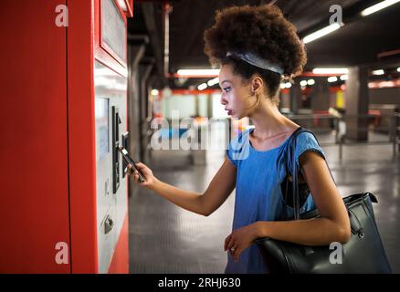 Side view of concentrated young mixed race female passenger with dark Afro hair using smartphone while paying for ticket from automatic machine in sub Stock Photo